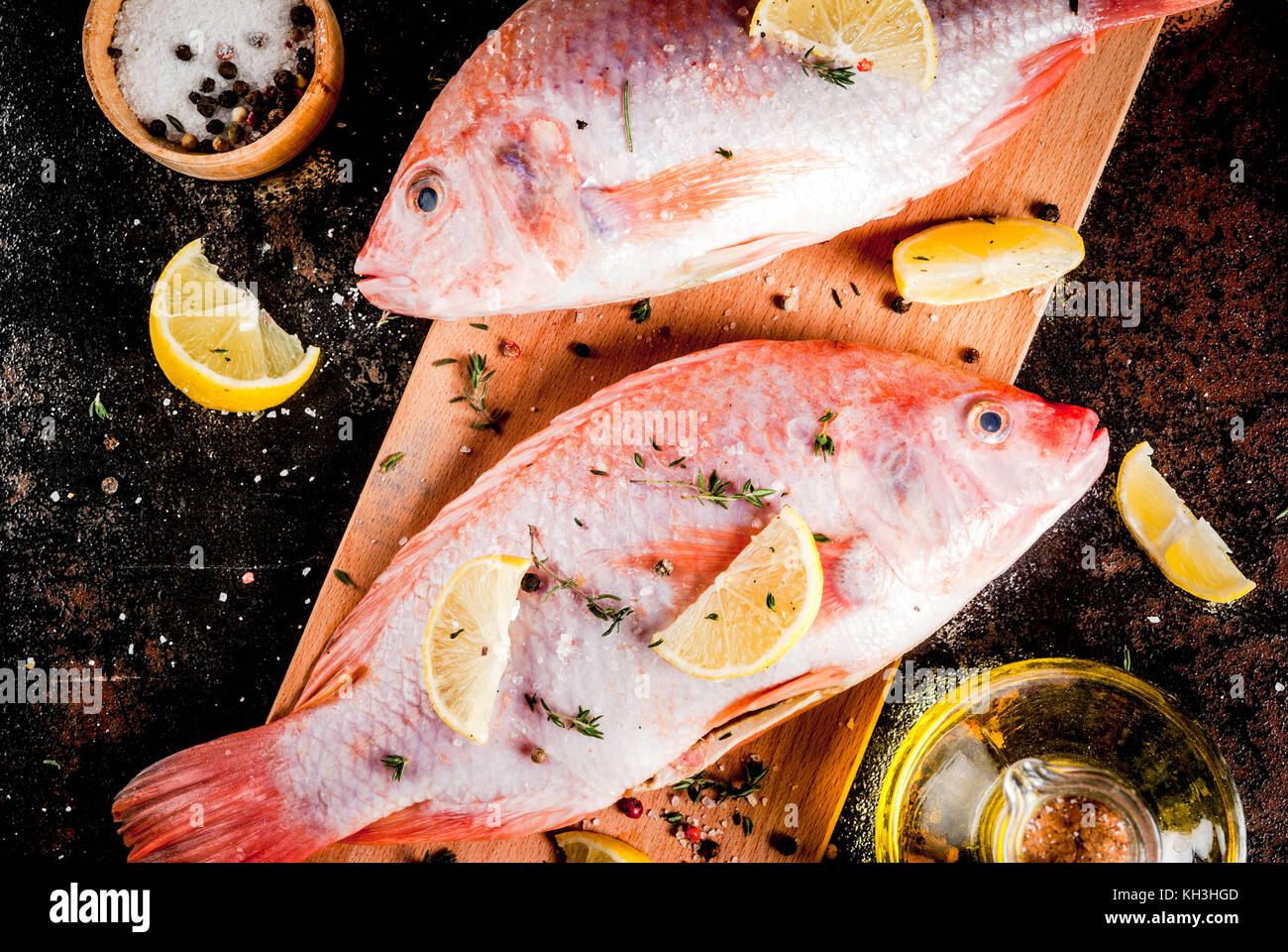 Frischen, rohen Fisch rosa Tilapia mit Gewürzen zum Kochen - Zitrone, Salz, Pfeffer, Kräuter, auf Schwarz rostiges Metall Tabelle, kopieren Raum, Ansicht von oben Stockfoto
