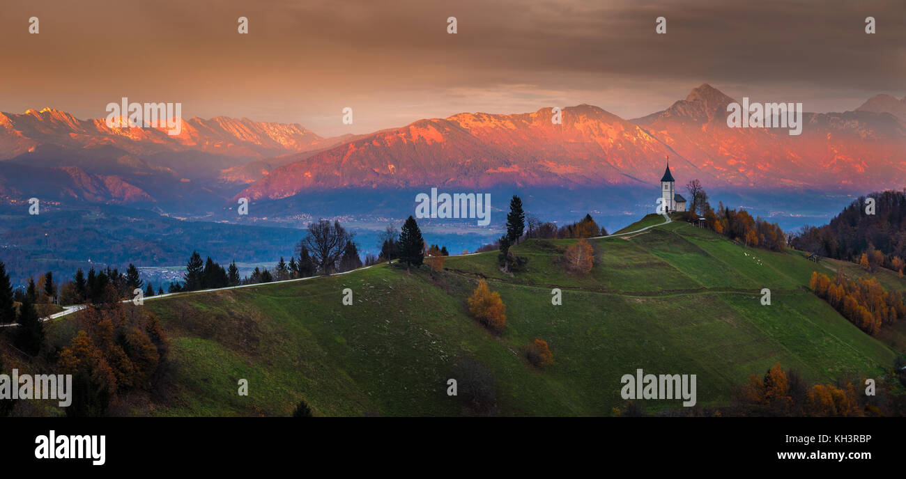 Jamnik, Slowenien - Schöner goldener Sonnenuntergang in der Jamnik St. Primoz Kirche. Julische Alpen im Hintergrund auf Panoramabild Stockfoto