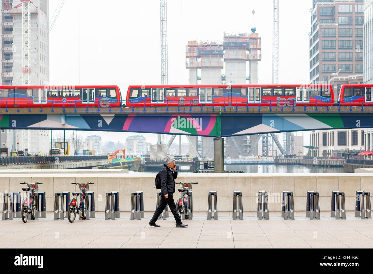 London, Großbritannien - 24 November, 2017 - Ein Geschäftsmann auf reuters Plaza in Canary Wharf mit DLR-Züge, und die Baustelle im Hintergrund Stockfoto