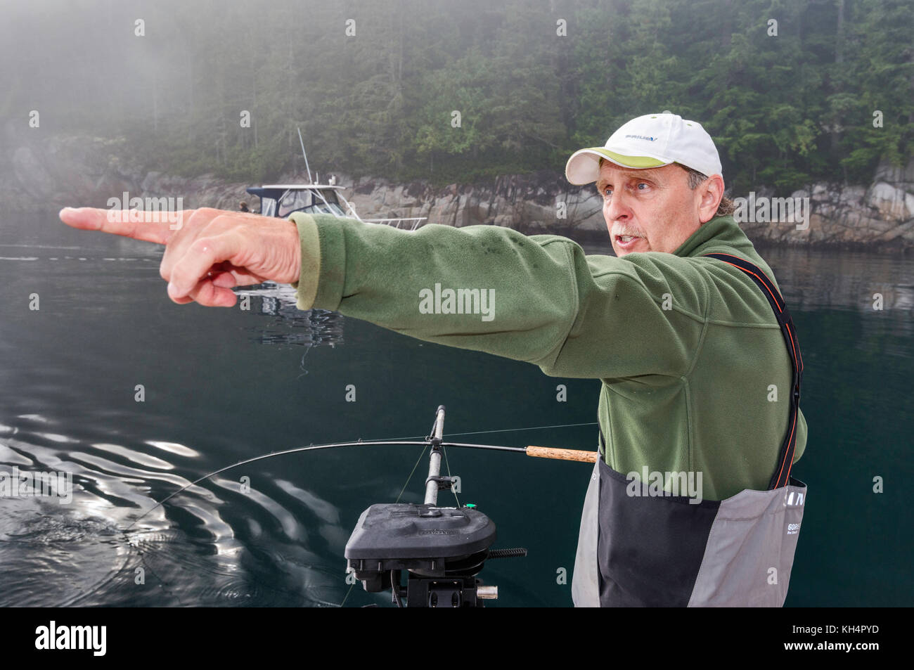 Erwachsener mittleren Alters mit Angelrute auf dem Boot, der darauf hinweist, dass ein anderes Boot vorbeikommt, nebeliger Morgen in der Johnstone Strait vor Vancouver Island, British Columbia Stockfoto