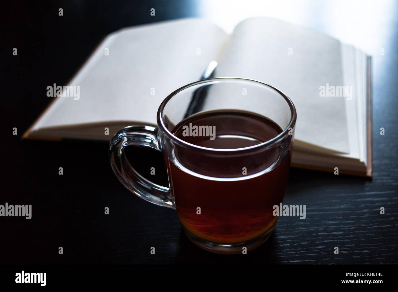 Ein Glas Becher mit Tee und einer Zitronenscheibe, Notebook mit leeren Seiten, schwarzen Kugelschreiber auf dunklen Schreibtisch Stockfoto