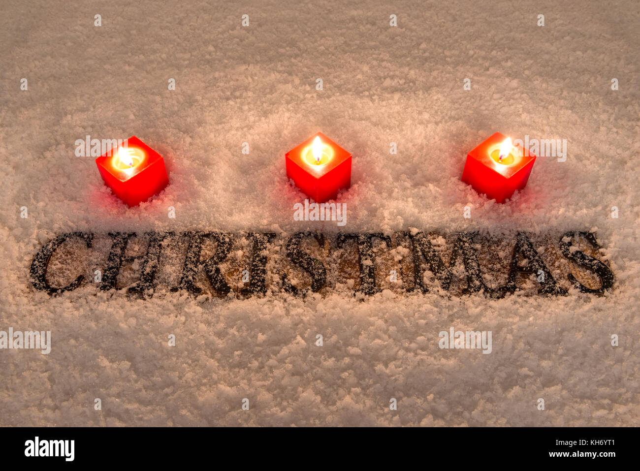 Das Wort Weihnachten in Holz- Buchstaben auf einem Schnee Hintergrund durch drei rote Kerzen beleuchtet. Stockfoto