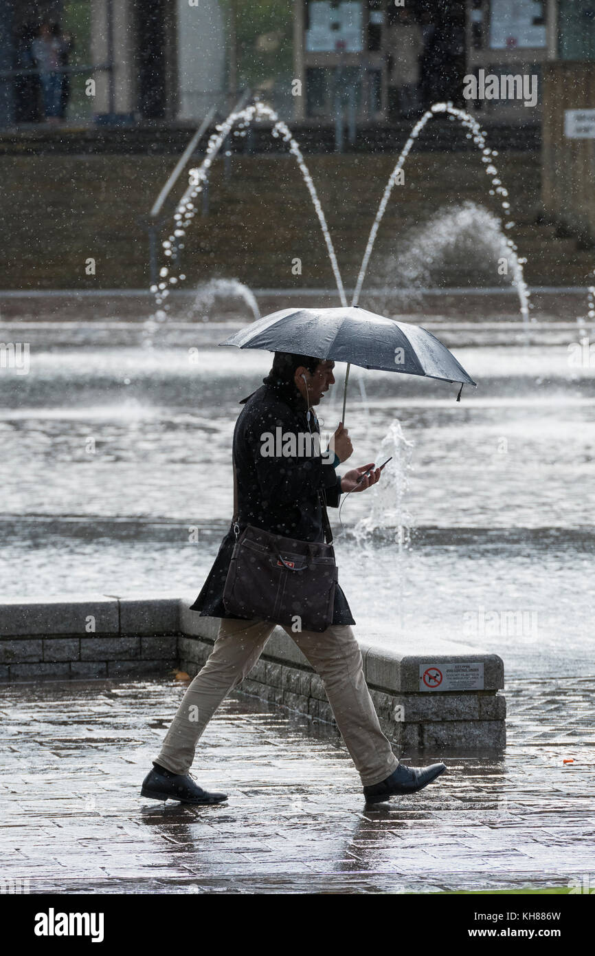 Mann in der schweren Regen, holding Dach & Phone, vorbei an den Spiegel Pool & Brunnen - Bradford City Park, West Yorkshire, England, UK. Stockfoto