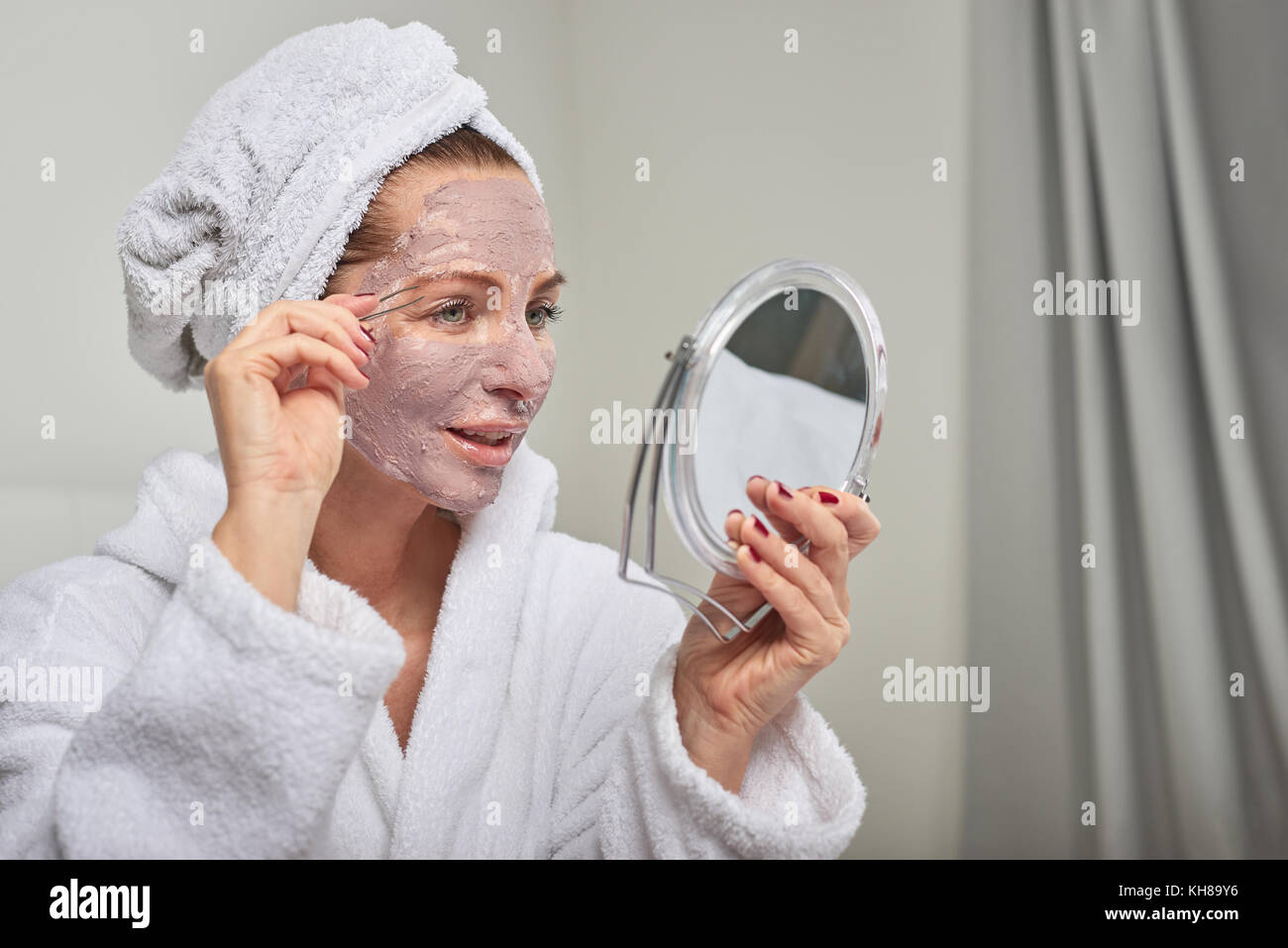 Attraktive Frau das Tragen einer Schutzmaske, Schönheitsbehandlungen, ein Spiegel, wie sie ihre Augenbrauen zupft mit Pinzette Stockfoto