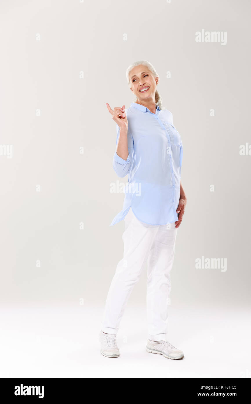 Full-length Portrait von spielerischen reife Frau in blauen Hemden und weißen Hosen, stehen und zeigt mit dem Finger, abgesehen suchen, auf weissem backgr isoliert Stockfoto