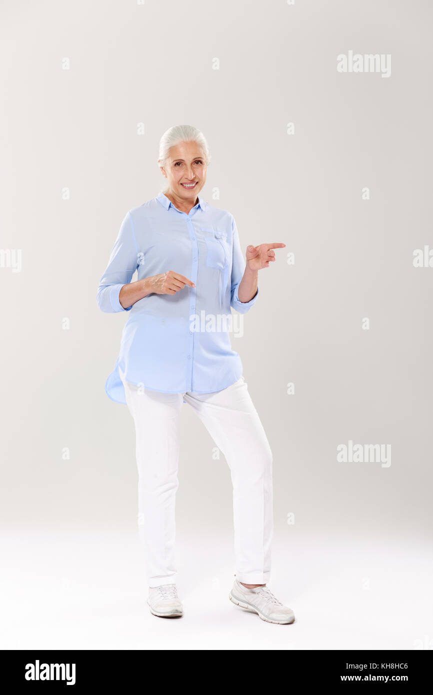 Full-length Portrait von gerne ältere grauhaarige Frau, stehen und zeigt mit Fingern, an der Kamera suchen, auf weißem Hintergrund Stockfoto