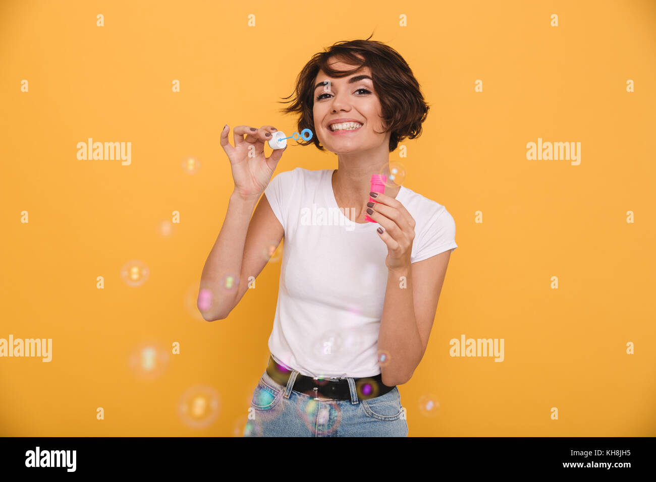 Porträt einer fröhlicher junger Mädchen bläst Seifenblasen im Stehen, und wenn man die Kamera auf gelben Hintergrund isoliert Stockfoto