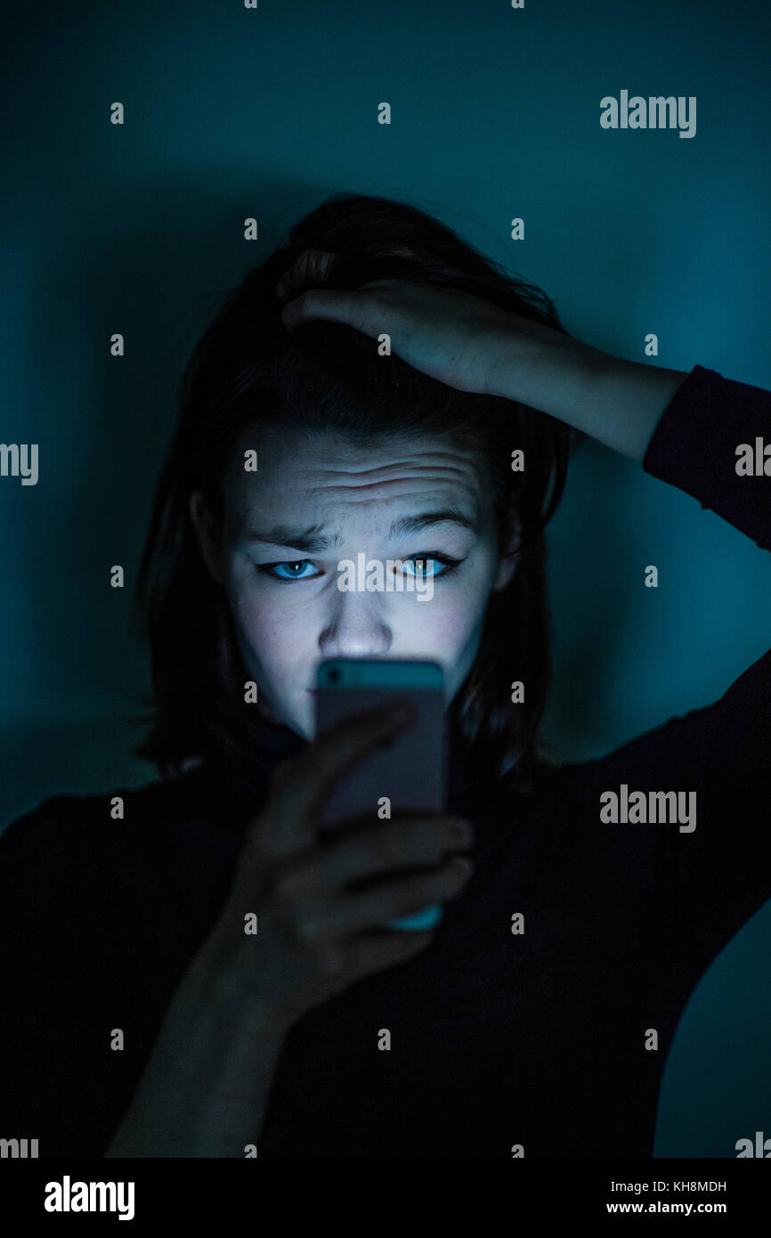 Cyber stalking: Eine junge Braunhaarige kaukasischen teenage Mädchen auf ihrem Mobiltelefon, Smartphone, Handy, besorgt suchen, verwirrt, besorgt durch den Inhalt der Botschaft, die sie ist zu sehen, in der Nacht, Großbritannien Stockfoto