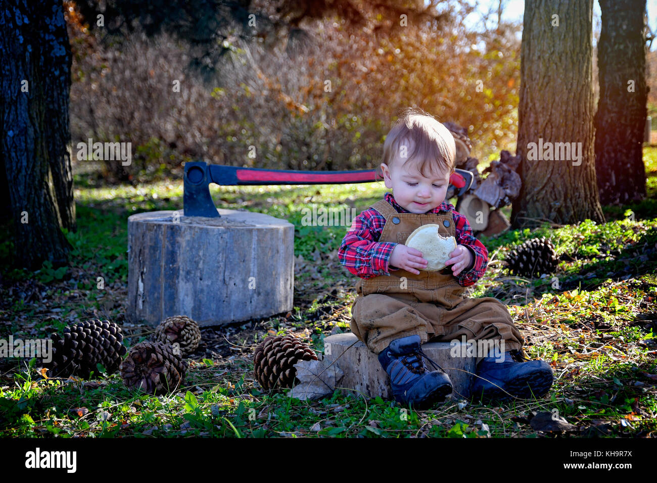 Baby Boy Holzfäller das Tragen von Stiefeln und Overalls sitzen Essen ein Sandwich ax im Hintergrund große Kiefernzapfen ländlichen Ländereinstellung Stockfoto