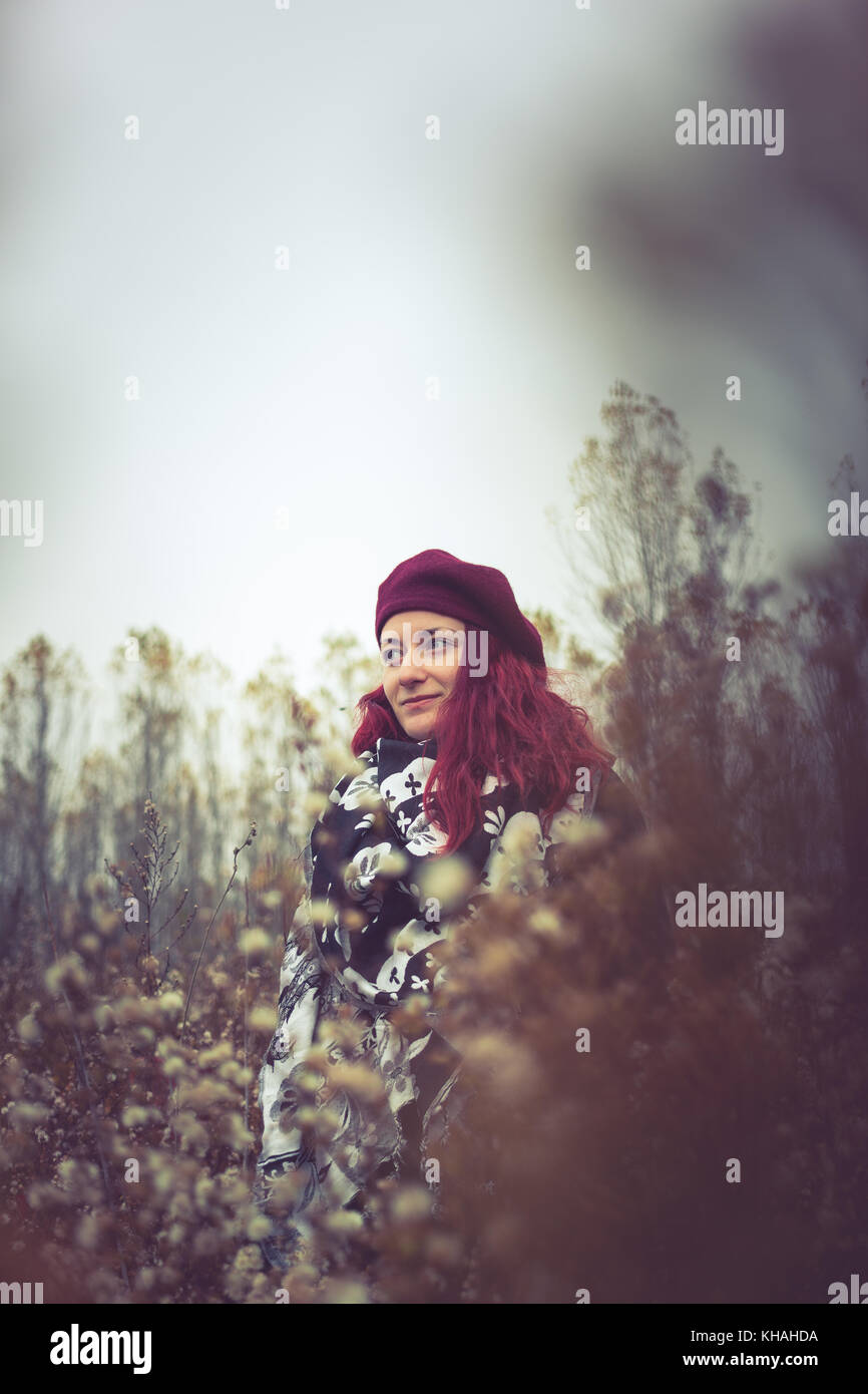 Junge Frau mit roten Haaren portrait stand mitten unter Blumen auf einem donauufer das Tragen der roten Baskenmütze und schwarzen und weißen Schal auf einer Entfernung Stockfoto