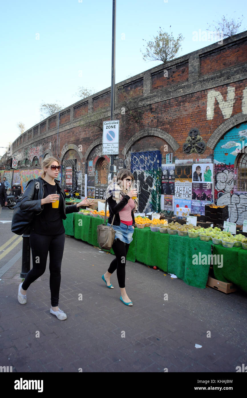 Junge Frauen, die Getränke zu Fuß vorbei an Obstständen in Shoreditch, London, England Stockfoto