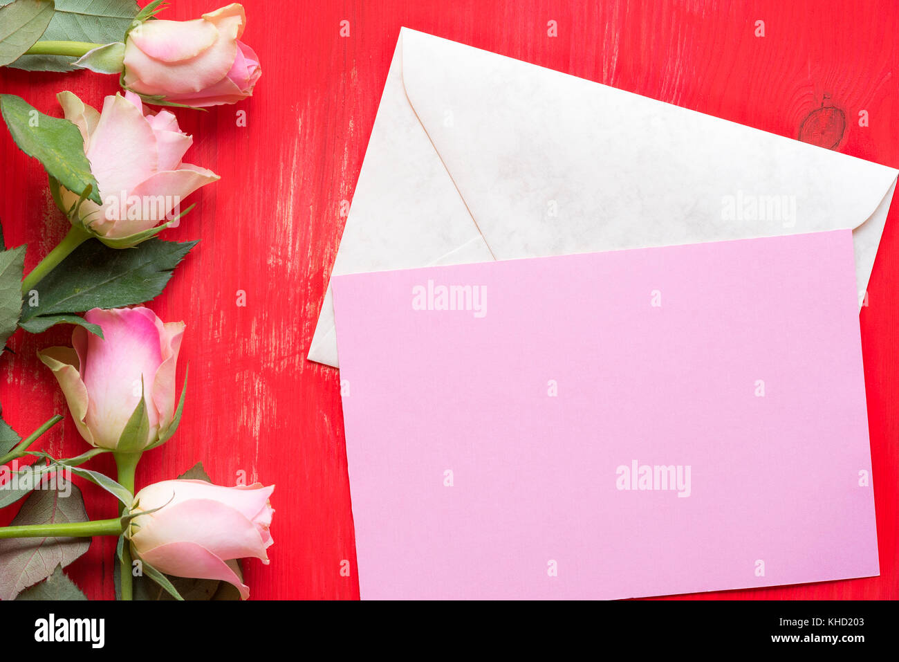 Grußkarte Idee mit einem rosa Papier in einem verschlossenen Umschlag, von Rosen auf einem roten Hintergrund Holz umgeben. ein Konzept für Muttertag, Valentinstag und Stockfoto