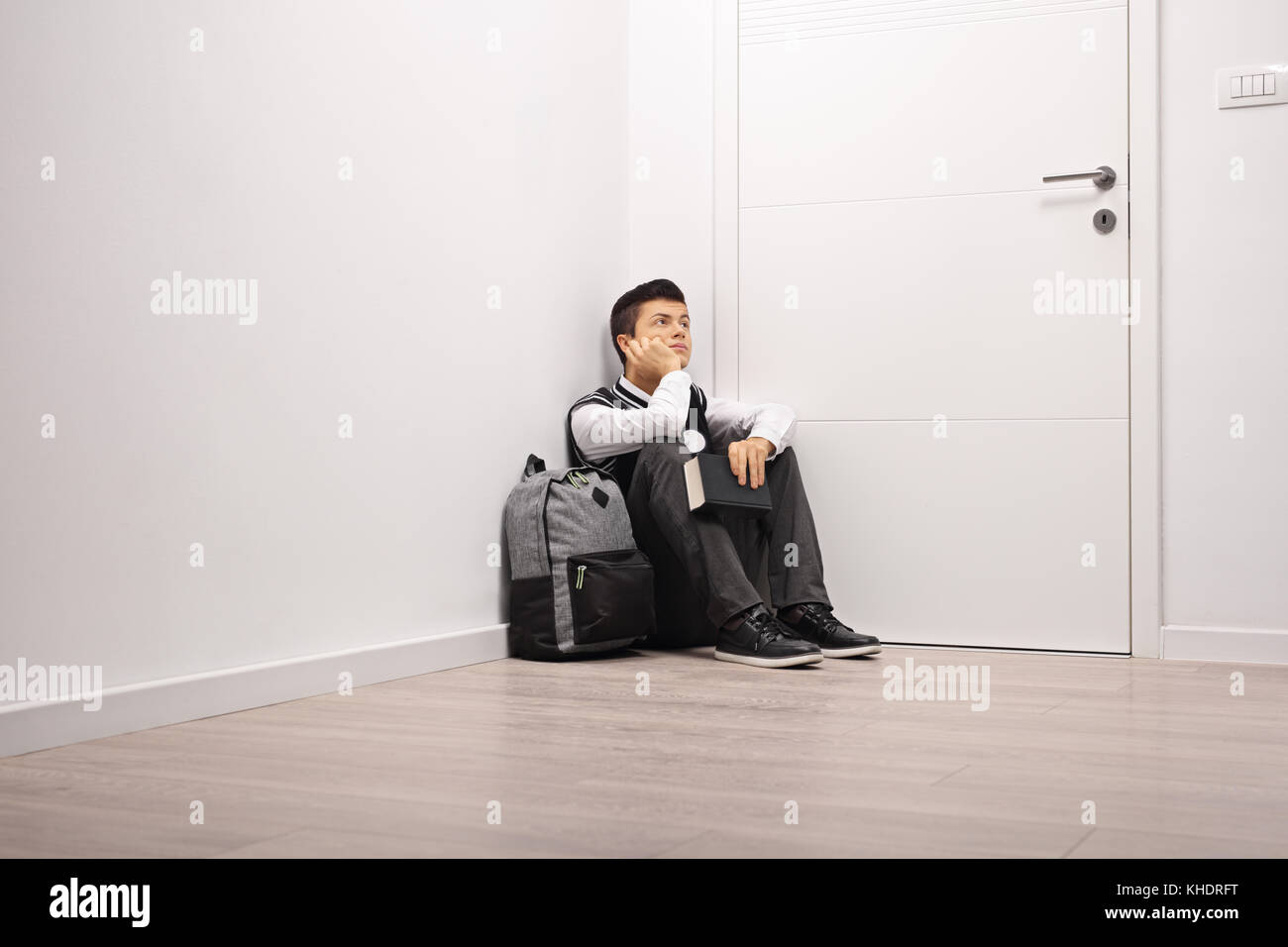 Nachdenklich teenage Student mit einem Rucksack und einem Buch in eine Ecke drinnen sitzen Stockfoto