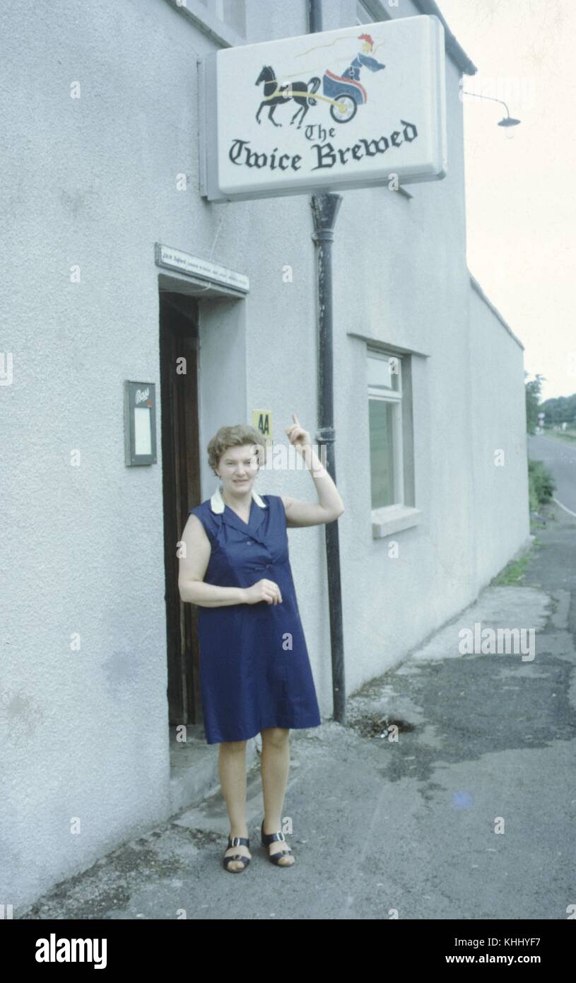 Ein Foto zeigt, dass eine Frau und deutete auf das Zeichen des gebraut Inn, ein öffentliches Haus und Hotel in Hexham, sie wird angezeigt, tragen ein ärmelloses blaues Kleid und steht neben dem einfachen grauen Putz Fassade des Business, Northumberland, Großbritannien, 1961. Stockfoto