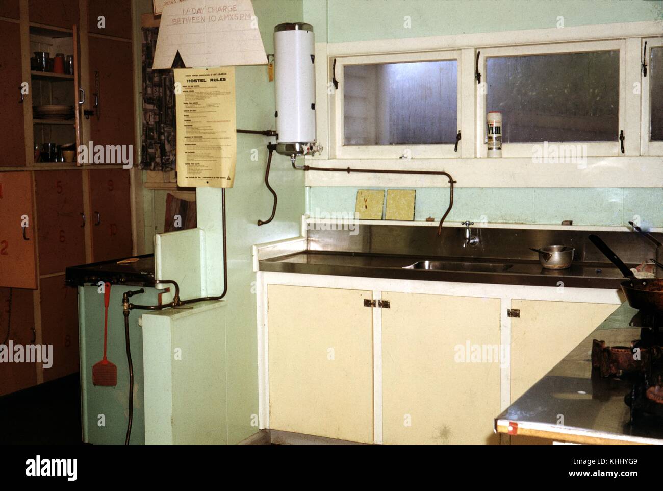 Ein Foto von einer Küche in einer Jugendherberge, die Zähler und Waschbecken aus Edelstahl abgedeckt werden, eine große Anzahl von Schränken kann im Hintergrund gegenüber einem Flat Top Grill gesehen werden, drei Fenster über der Spüle und Arbeitsplatte, Neuseeland, 1961 befinden. Stockfoto