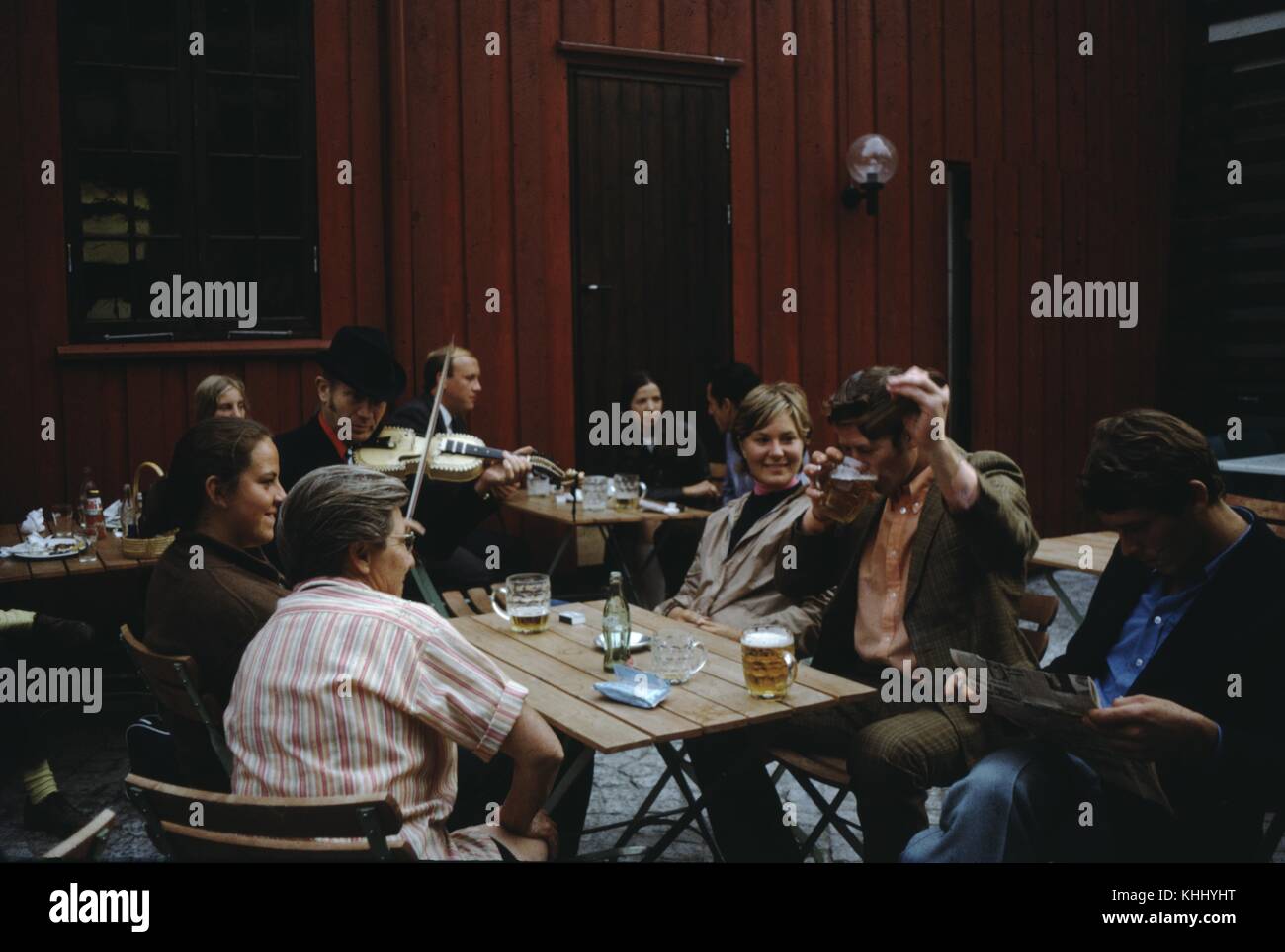 Ein Foto, das eine Familie um einen Tisch in einem Pub, der Vater ist unterhaltsam die Frauen der Familie durch das Trinken von Bier kräftig, der Sohn ein Papier liest, während ein Mann spielt eine Violine Musik dazu spielt, andere Gruppen von Menschen können im Hintergrund der Terrasse, 1961 gesehen werden. Stockfoto