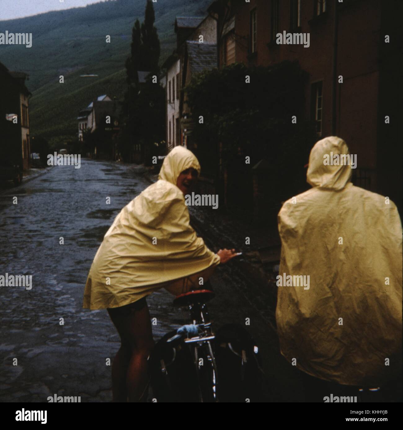 Zwei weibliche Radfahrer im Regen an einem bewölkten Tag in einer kleinen europäischen Stadt gefangen, Lachen, Kriechen auf gelb Regen Slickers, und zurück klettern an Bord ihrer Fahrräder, 1960. Stockfoto
