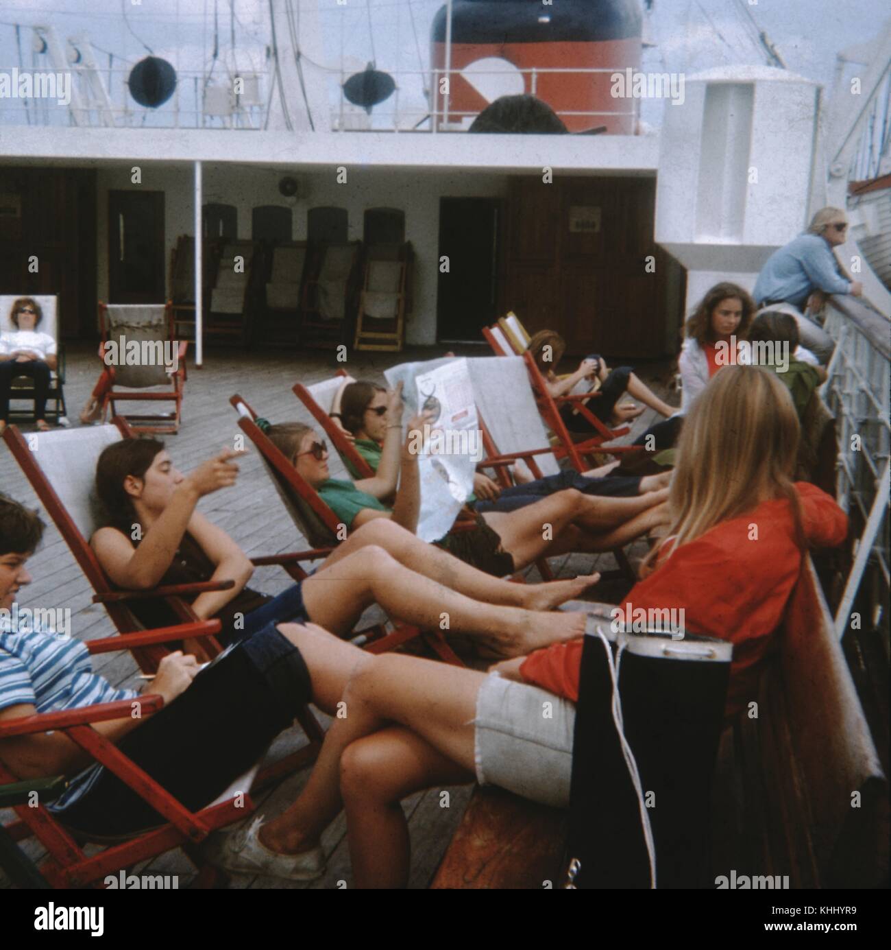 Familie, die sich entspannt und Zeitungen liest, während sie auf Liegestühlen auf dem oberen Deck eines Dampfschiffes ausruhen (1961). Stockfoto