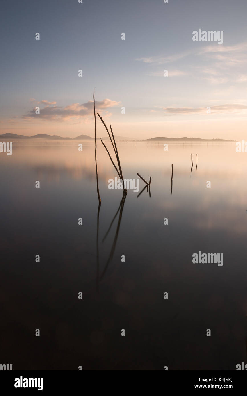 Perfekte Reflexionen einer Abenddämmerung auf dem See, mit den Wolken und Hügel auf dem Wasser spiegelt, weiche und warme Farben, und einige Holzpfähle aus dem Wasser kommt Stockfoto