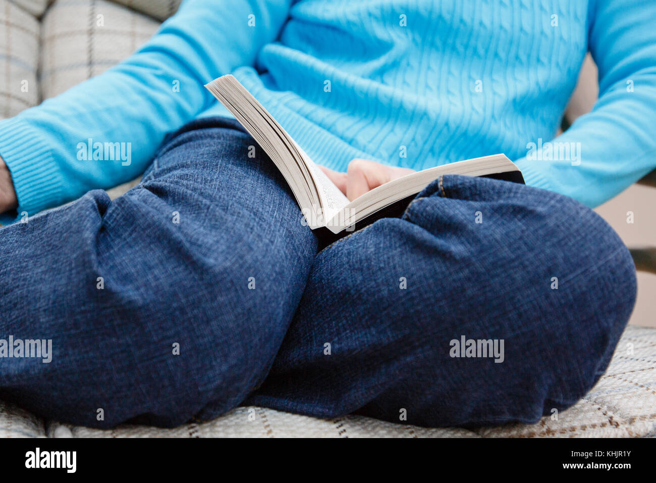 Eine Seniorin, die eine blaue Denim-Jeans trägt, sitzt auf einer Stuhllesung, und ein Buch ruht auf ihren Knien, das sich zuhause entspannt. England Großbritannien Stockfoto