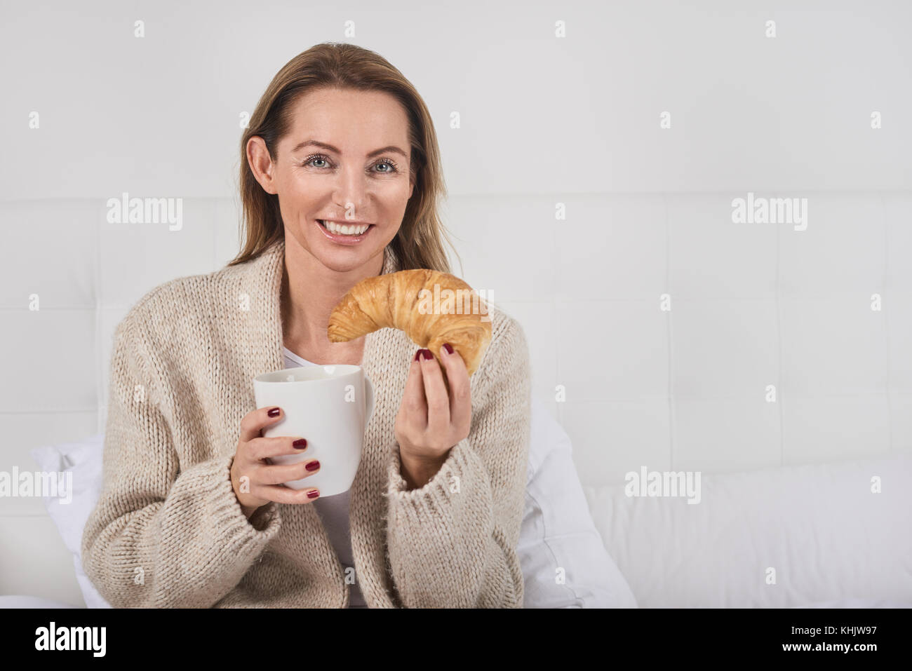 Porträt einer schönen Frau lächelnd während Sie frühstücken im Bett mit einer Tasse Kaffee oder Milch und einem köstlichen französischen Croissant Stockfoto