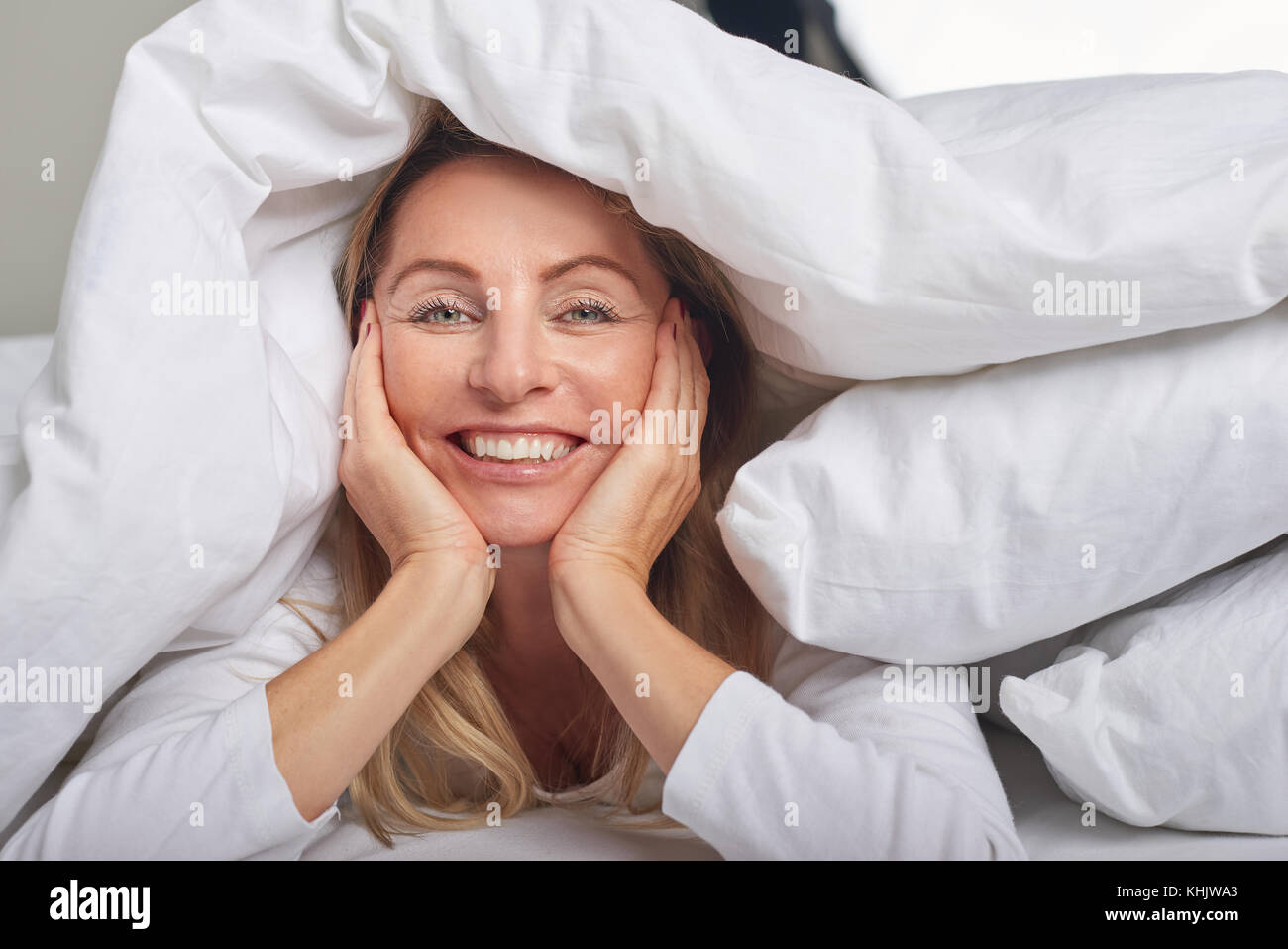 Schöne Frau mittleren Alters unter weißen Laken auf Kamera mit einem glücklichen Lächeln Stockfoto