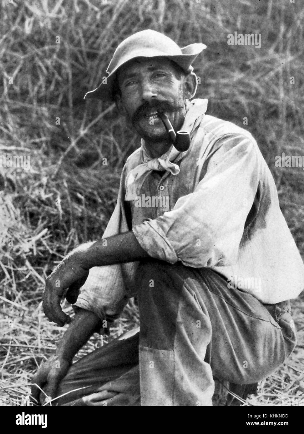 Ein Foto, das ein ganzes Porträt eines Landarbeiters zeigt. Er ist niedergeknickt und raucht eine Pfeife unter trockenem Gras. Er trägt einfache Kleidung, die von Schmutz und Schweiß bedeckt ist, aus seiner Arbeit, Camargue, Frankreich, Juli 1922. Stockfoto