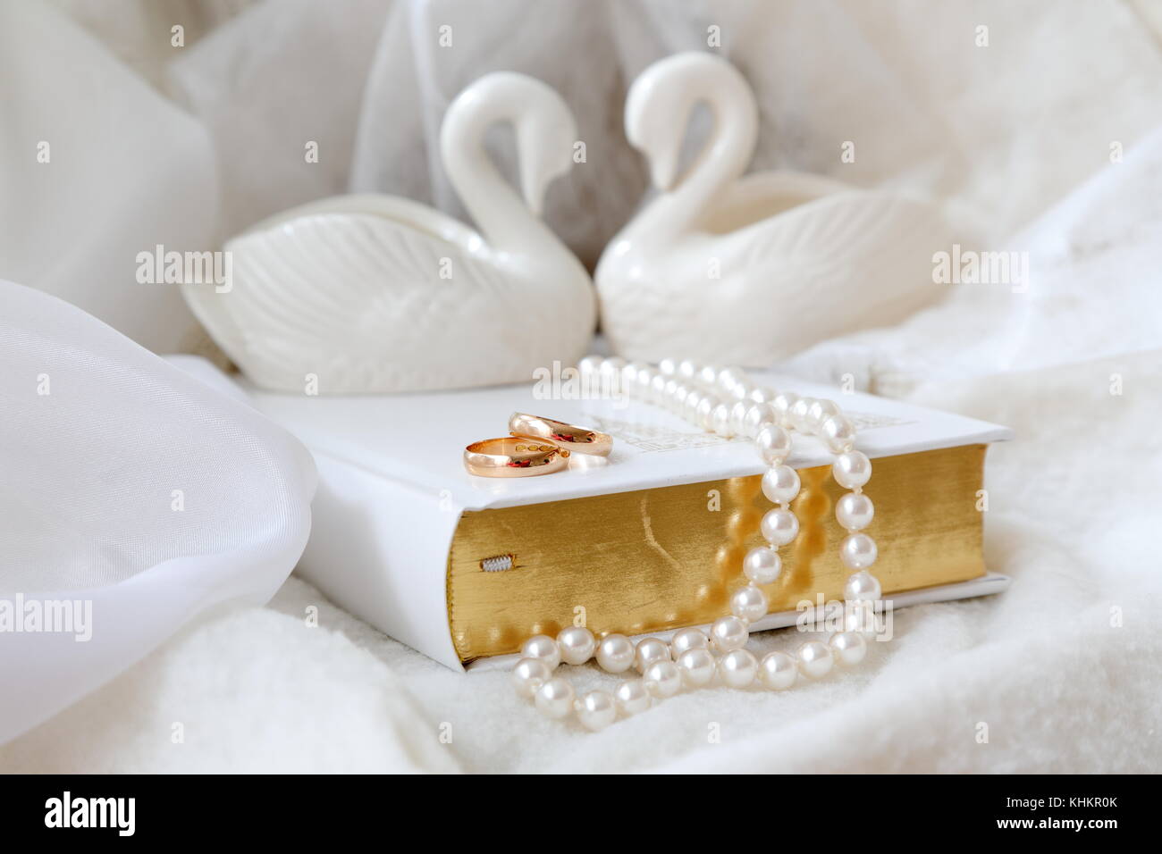 Zwei goldene Eheringe auf der weißen Bibel. Auch weiße Perlen und zwei weiße Porzellanschwäne. Weißer Hintergrund. Stockfoto