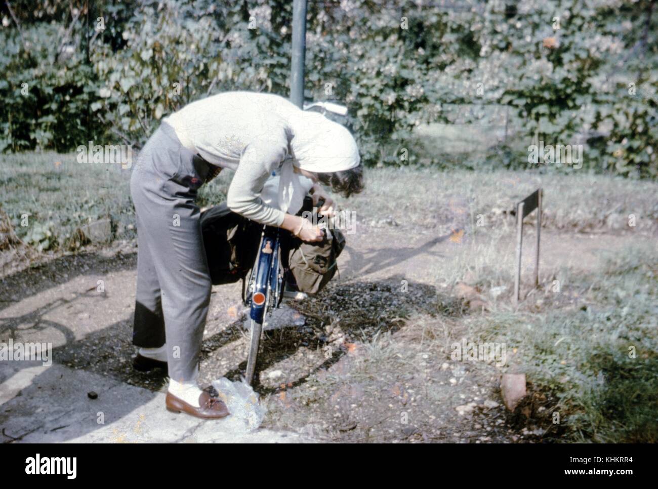Ein Foto von einer Frau mit ihrem Fahrrad, sie versucht, ein Objekt aus der Satteltasche abzurufen, sie trägt graue Hose mit braun Slipper und ein weißes T-Shirt mit einem weißen Kopftuch, das Fahrrad ist neben einem kleinen Schmutz weg, 1965 geparkt. Stockfoto