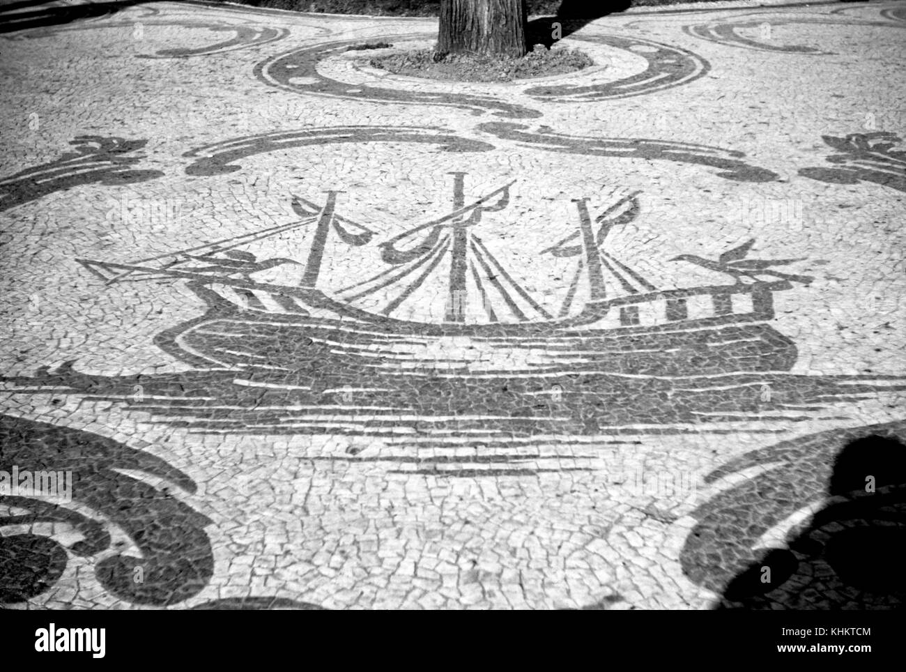 Eine Fotografie eines Mosaiks aus Fliesen im Freien, das sich um einen Baum wickelt, der Mosaikhintergrund ist weiße Fliese, alle Details sind in einem dunkelgrauen, ein Segelschiff mit zwei großen Vögeln an jedem Ende, es ist auf dem Wasser zu sehen und andere Designs, die Wind oder Wellen darstellen, sind ebenfalls zu sehen, 1965. Stockfoto