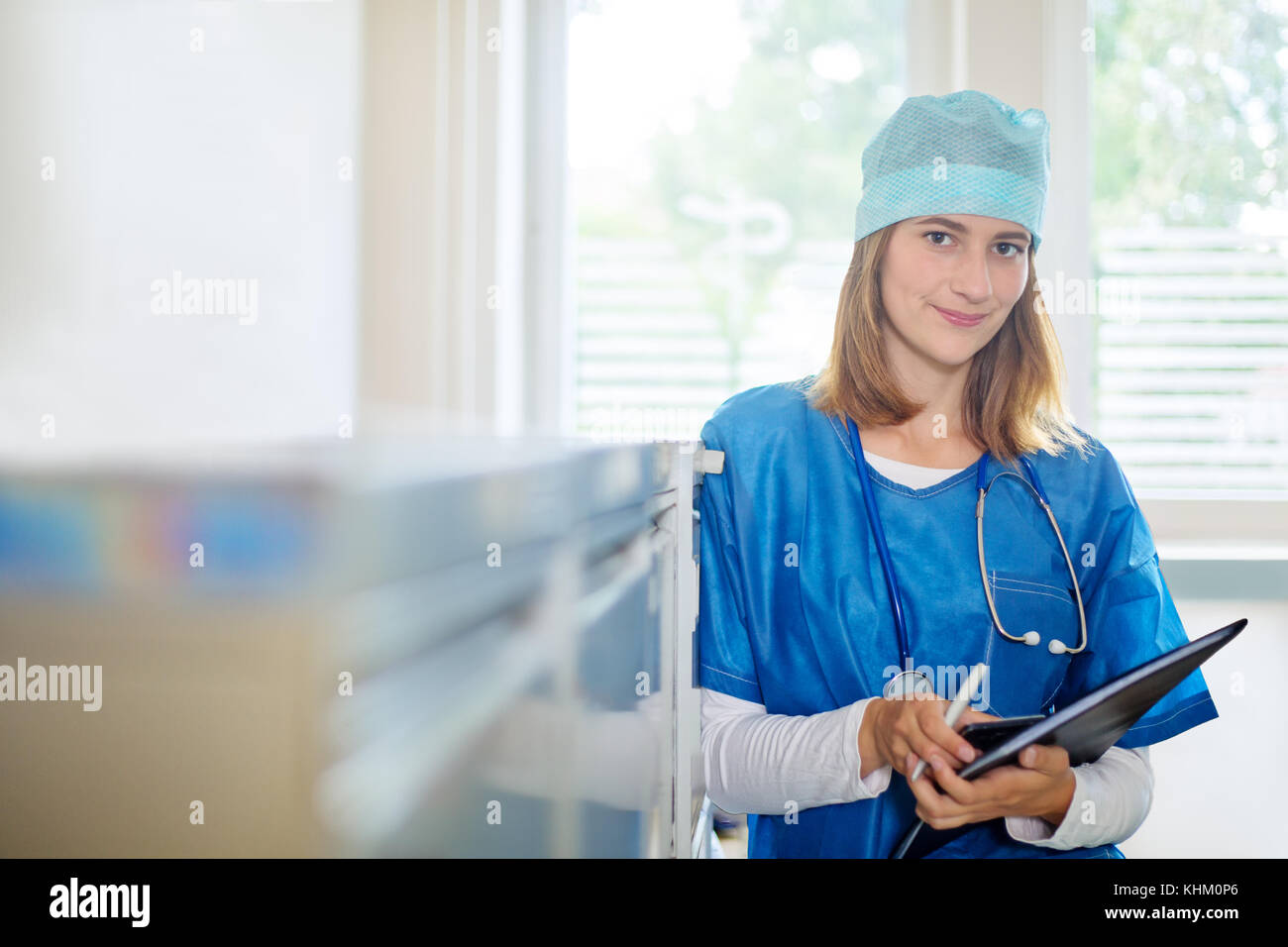 Junge weibliche Arzt in einer blauen Uniform steht drinnen in einer Klinik, mit Handy, Kugelschreiber und Ordner, in Richtung Aktenschrank lehnend, lo Stockfoto