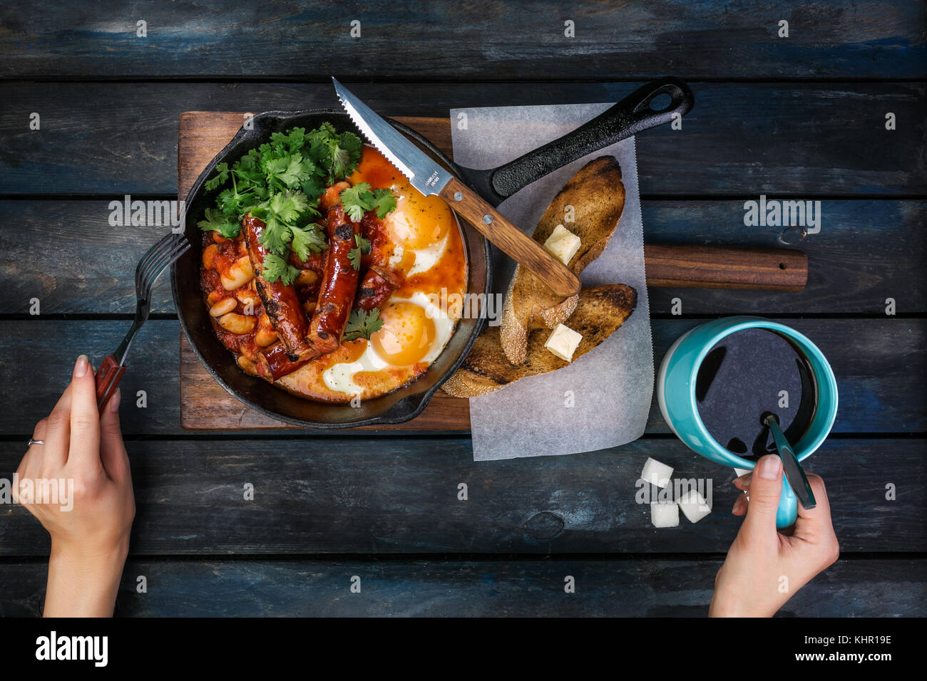 Frühstück in einer heißen Pfanne mit Spiegelei, Würstchen, Bohnen, Grün und Toasts. Frauen Hände mit Kaffee und Besteck. Auf einem farbigen Holz- Bac Stockfoto