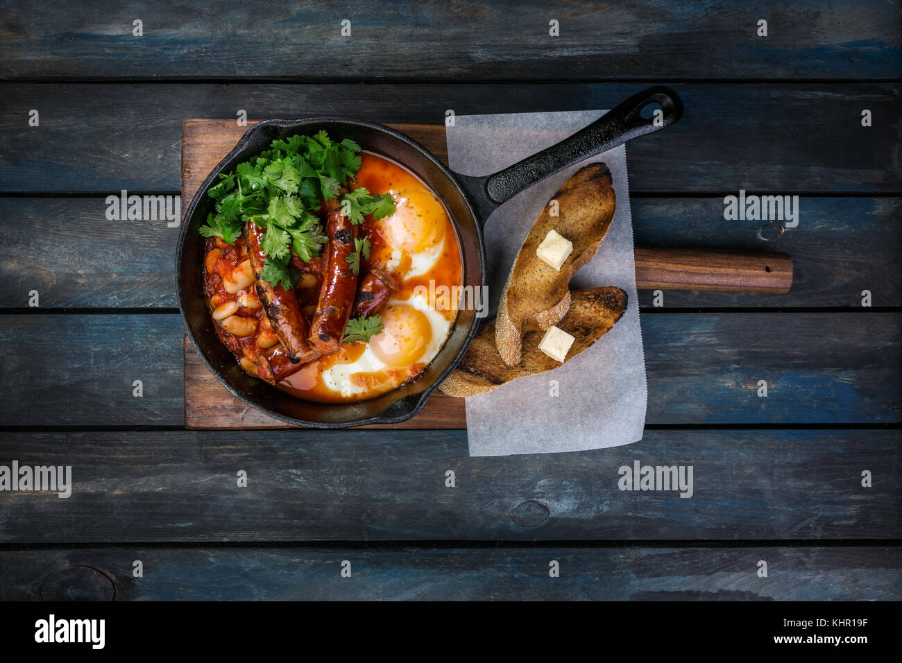 Frühstück in einer heißen Pfanne mit Spiegelei, Würstchen, Bohnen, Grün und Toasts. Auf einem farbigen Hintergrund Holz. nach oben anzeigen. Stockfoto