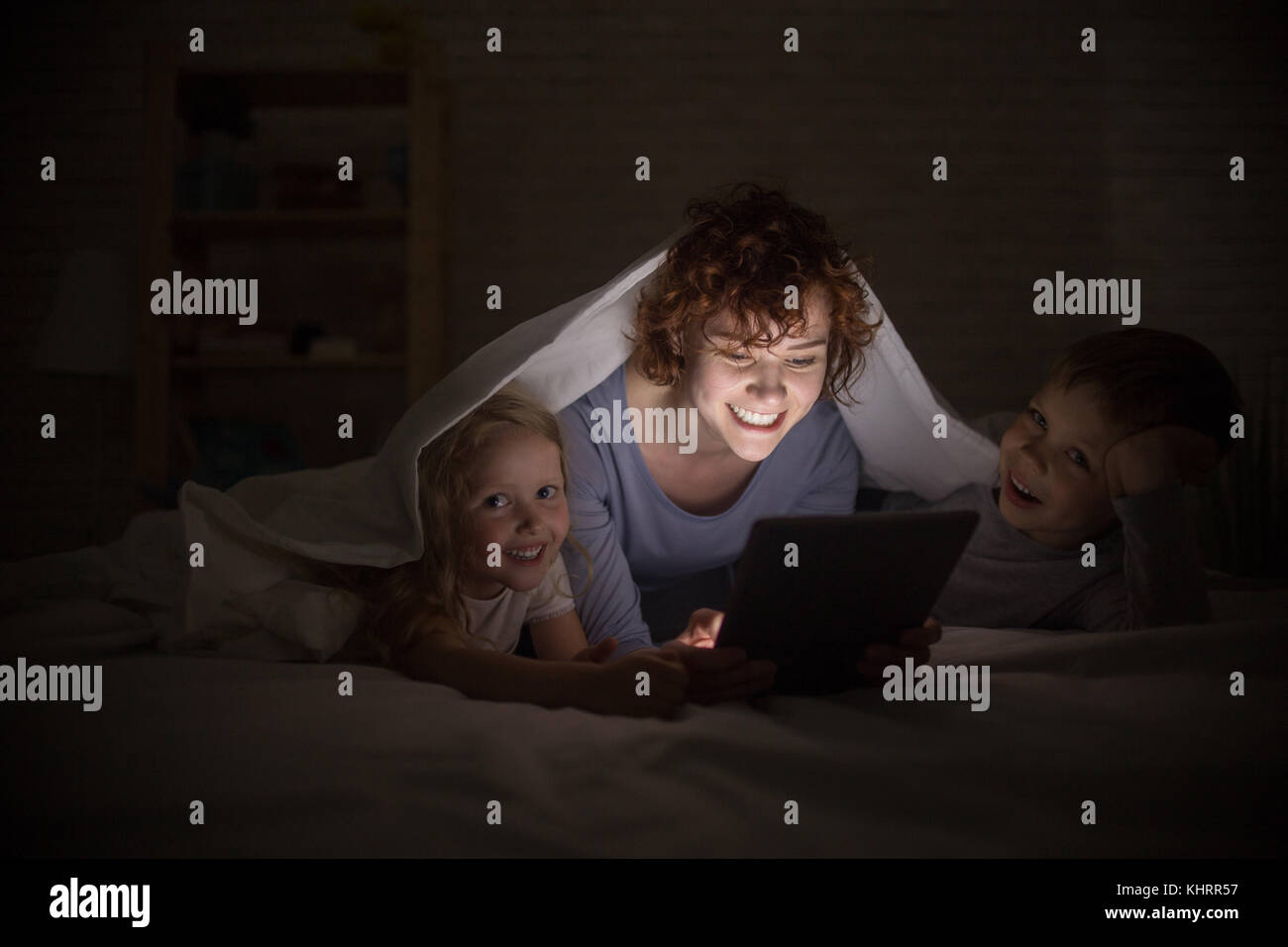 Porträt der glückliche junge Mutter mit zwei Kindern lesen Geschichten im Bett, versteckt unter Vertrag und e-book Stockfoto
