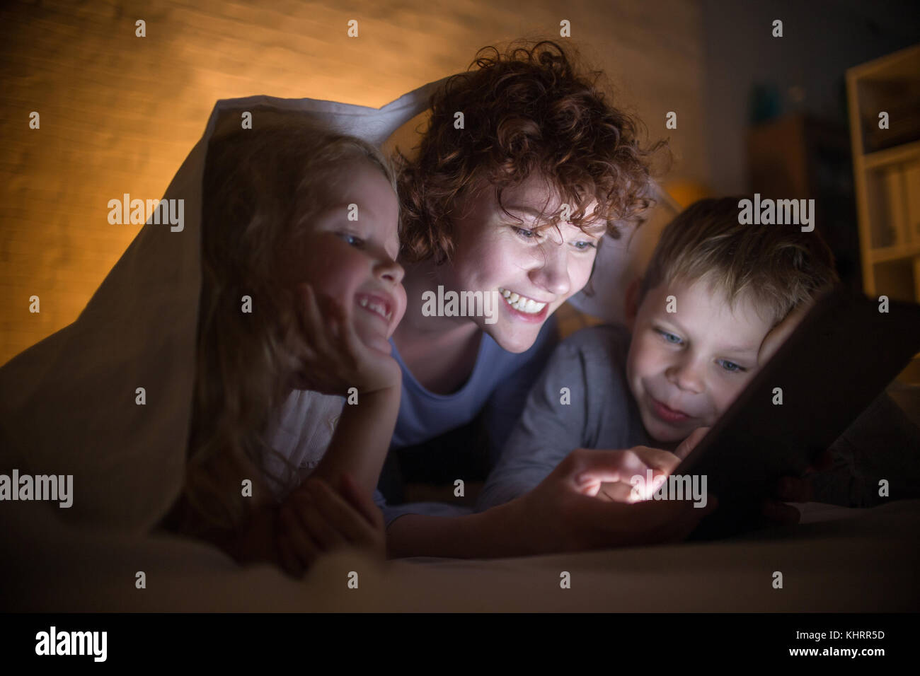 Porträt der glückliche junge Mutter mit zwei Kindern lesen Geschichten im Bett, versteckt unter Decke und mit Tablet Stockfoto