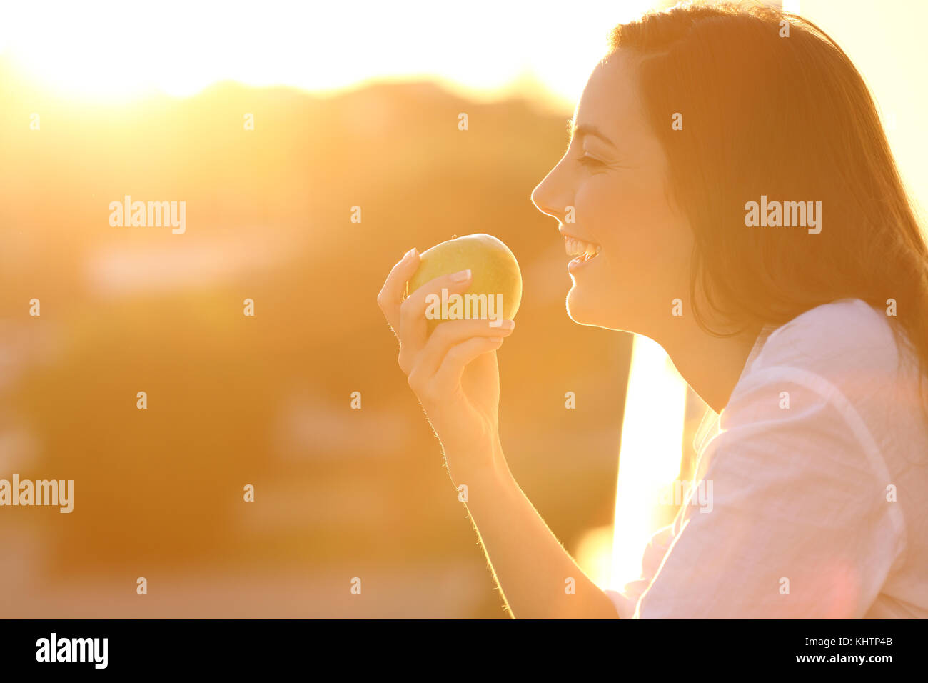 Seitenansicht Hintergrundlicht Porträt einer glücklichen Frau essen ein Apfel im Haus Balkon bei Sonnenuntergang Stockfoto