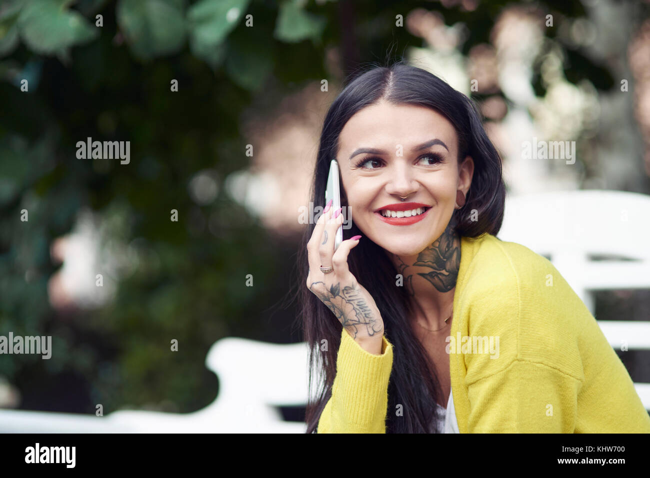 Junge Frau sitzt im Freien, mit Smartphone, lächelnd, Tattoos auf Hals und Hand Stockfoto