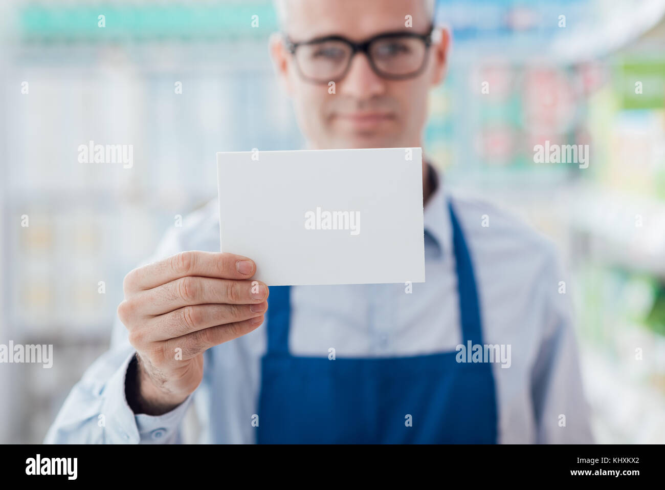 Lächelnd Supermarkt Arbeiter Holding ein leeres weißes Schild und Kamera, Regale auf dem Hintergrund Stockfoto