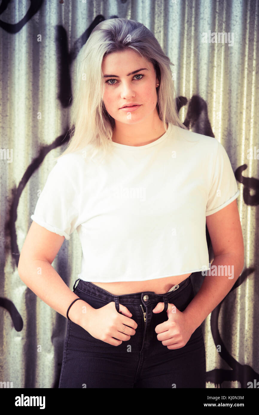 Jugendmädchen Modellierung in Großbritannien: Eine junge 16-jährige Teenager Modell, Moody, stehend mit Ihren Daumen in ihren Gürtelschlaufen, trug ein weißes T-Shirt, posiert vor einem Graffiti Wellblech metall Wand, uk abgedeckt Stockfoto