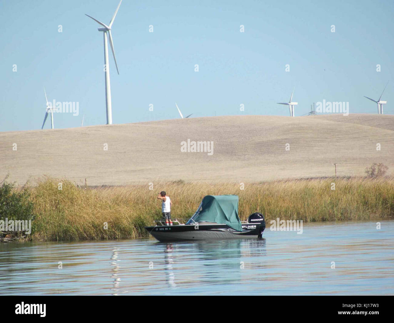 Ein Foto, das Männer in einem Boot auf einem Fluss einfängt, mit einem modernen Windpark im Hintergrund, der traditionelle und erneuerbare Energielandschaften vereint. Stockfoto