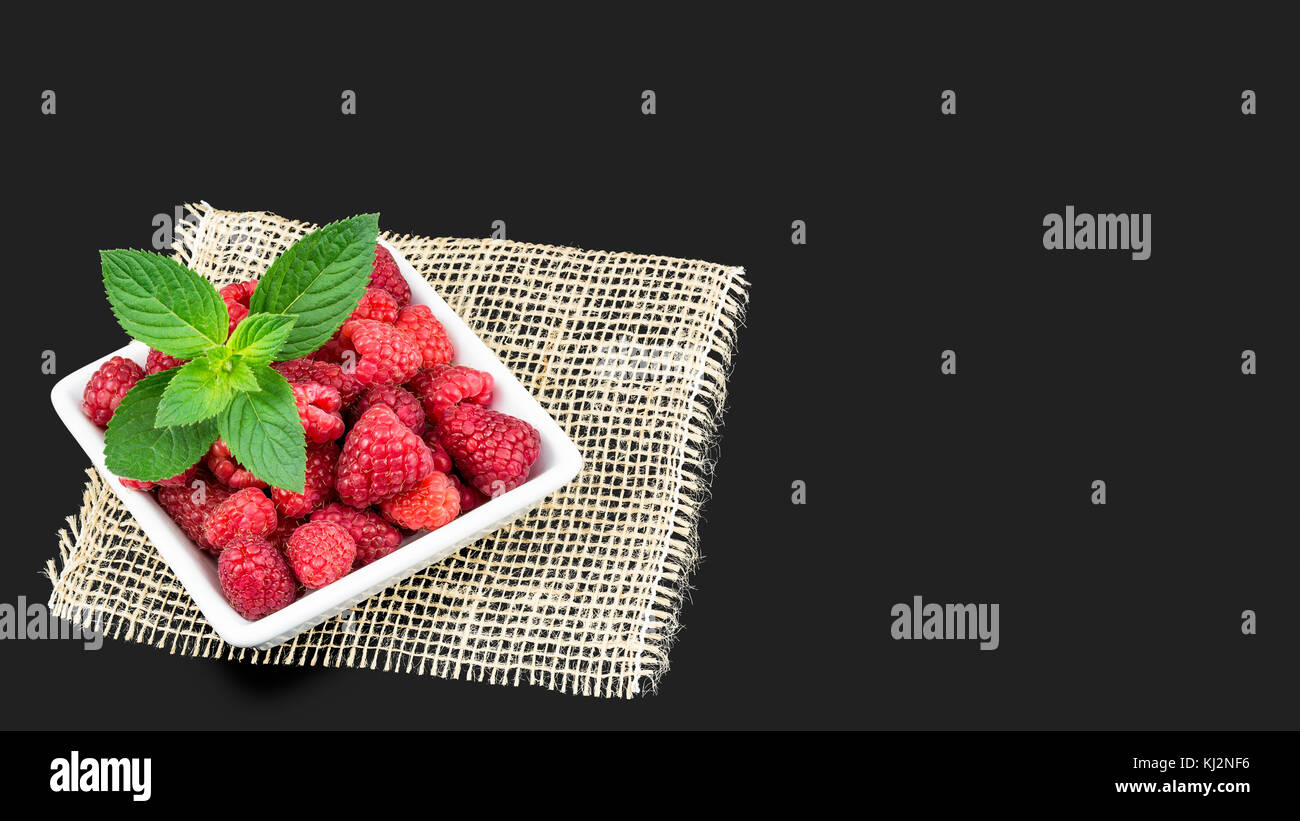 Köstliche frische Himbeeren auf einem dunkelgrauen Hintergrund. saftige rote Früchte im weißen Quadrat Schüssel mit Pfefferminze und dekorative Matte. Stockfoto