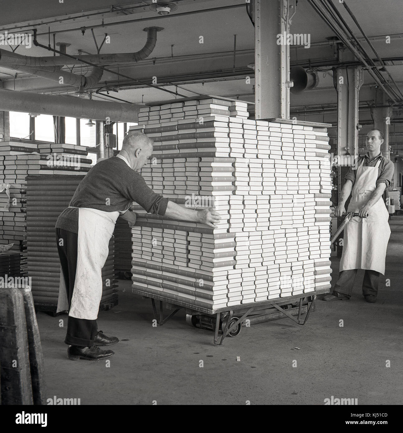 1950, historische, zwei männliche Arbeitnehmer Schürzen tragen in einer industriellen Anlage oder Lager, eine große und schwere Last von gebundene Bücher auf einem Transportwagen, England, UK. Stockfoto