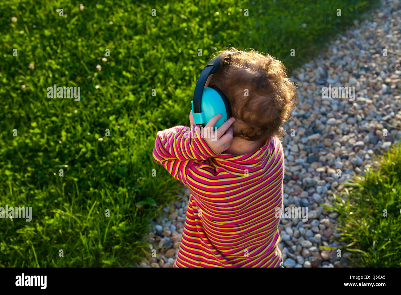 Kleinkind rothaarige Mädchen steht auf dem Rasen bei Sonnenuntergang mit dem Rücken zur Kamera Musik mit großen Kopfhörer Stockfoto