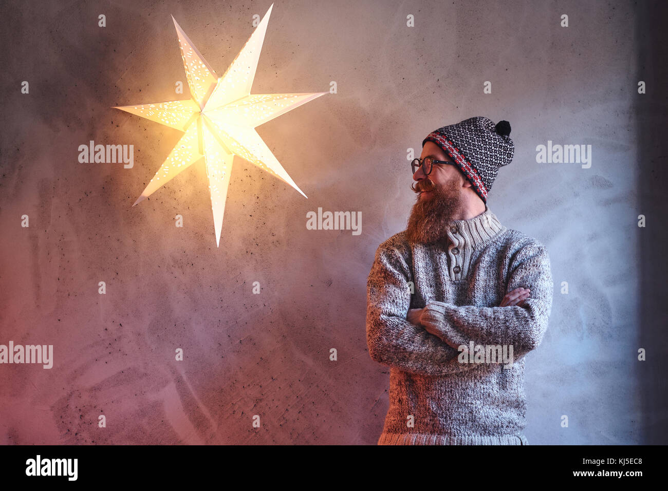 Bärtige Mann in einem warmen Pullover und eine Mütze über die Mauer mit einem dekorativen Stern Lampe gekleidet. Stockfoto