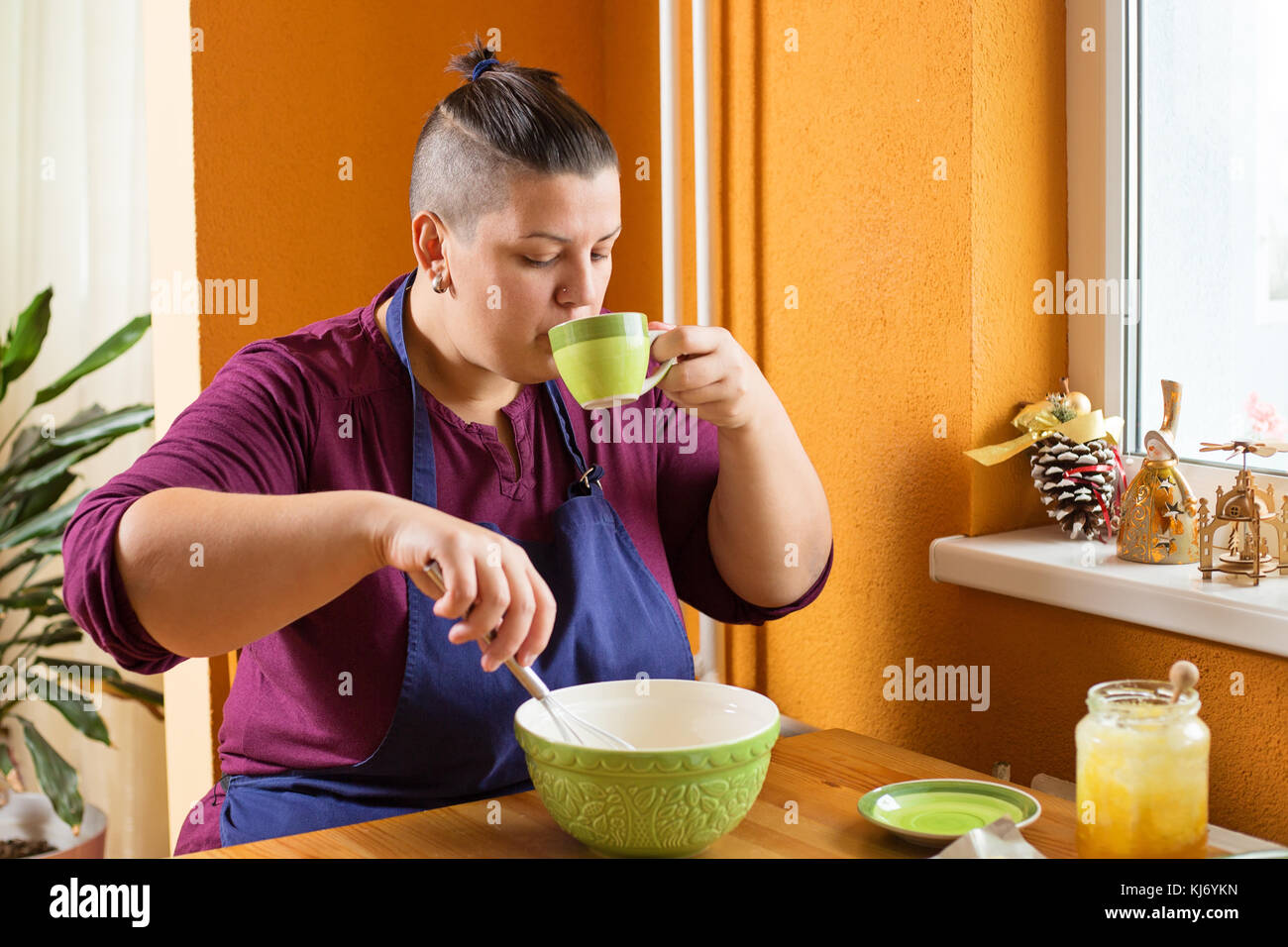 Schöne junge Frau mit kurzen Haaren sitzen in der Küche hinter einer hölzernen Tisch, mischen Sie die Zutaten in die Schüssel, während Sie einen Grünen Tee aus Gree Stockfoto