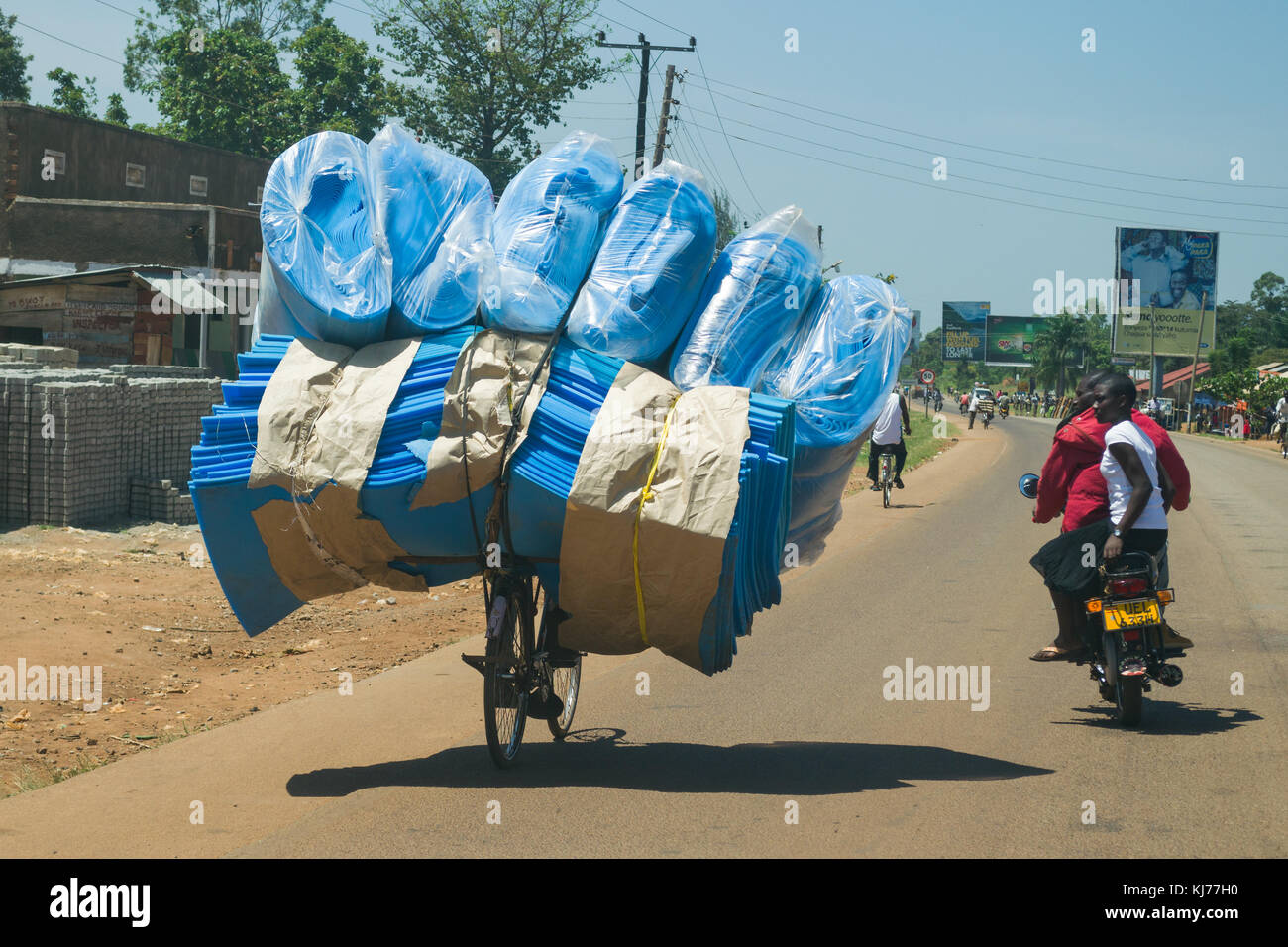 Eine bicycist schwer beladen mit blauen Schaumplatten Fahrten entlang einer Straße durch eine geschäftige Stadt, Busia, Uganda, Ostafrika Stockfoto