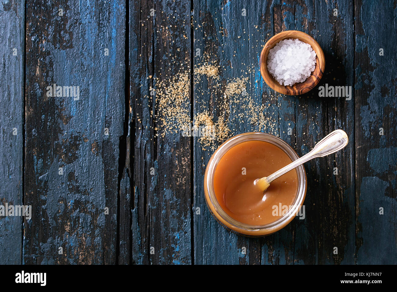 Glas Glas hausgemachten gesalzen Karamellsauce mit Löffel, brauner Zucker und einer Schale mit Salz. Über alte dunkelblaue Holz- Hintergrund. Ansicht von oben mit Platz Stockfoto