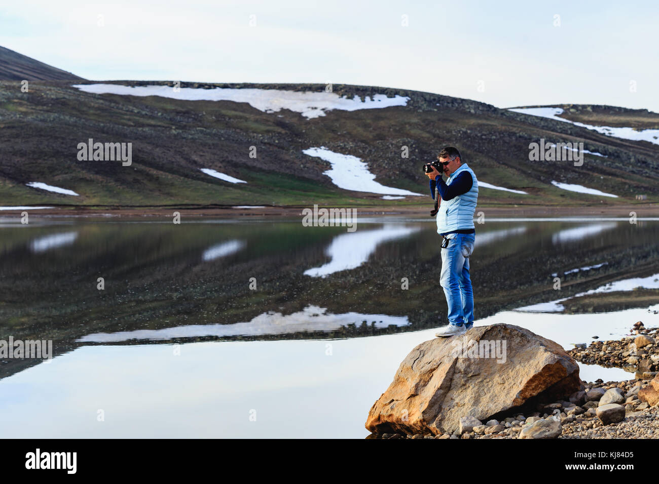 Horizontale Schuß von landscape photographer stehen auf Stein und trägt blaue Jeans und blass blaue Weste fotografieren Bergsee mit schmelzender Schnee Stockfoto