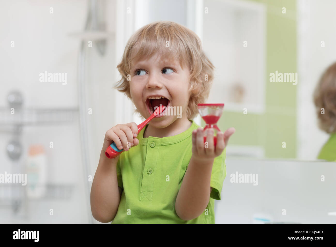 Glückliches Kind Zähneputzen in der Nähe der Spiegel im Bad. Er dauerhafte Überwachung der Reinigung mit Sanduhr. Stockfoto