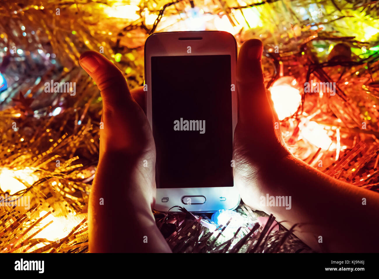 Kind ein Geschenk in Form einer weißen Smartphone unter dem Baum für das neue Jahr. Die Hand am Telefon auf dem Hintergrund der Weihnachtsbeleuchtung Stockfoto